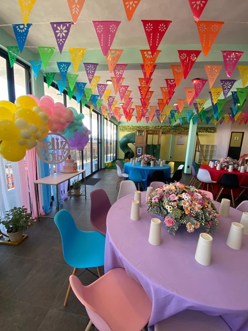 Salón Perinolita montado con mesas de colores, papel picado, arco de globos y área de juegos al fondo